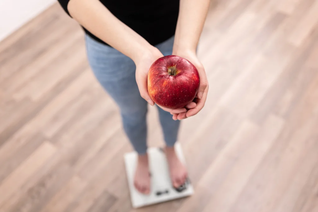 Persona de pie sobre una báscula sosteniendo una manzana, representando una alimentación saludable y el éxito del Balón Gástrico Ingerible en Zipaquirá.