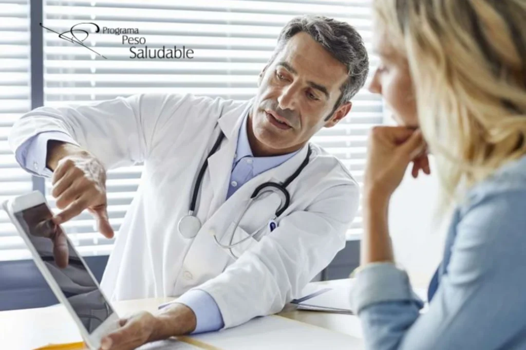 Médico con bata blanca y estetoscopio mostrando información en una tableta digital a una paciente durante una consulta del Programa Peso Saludable.